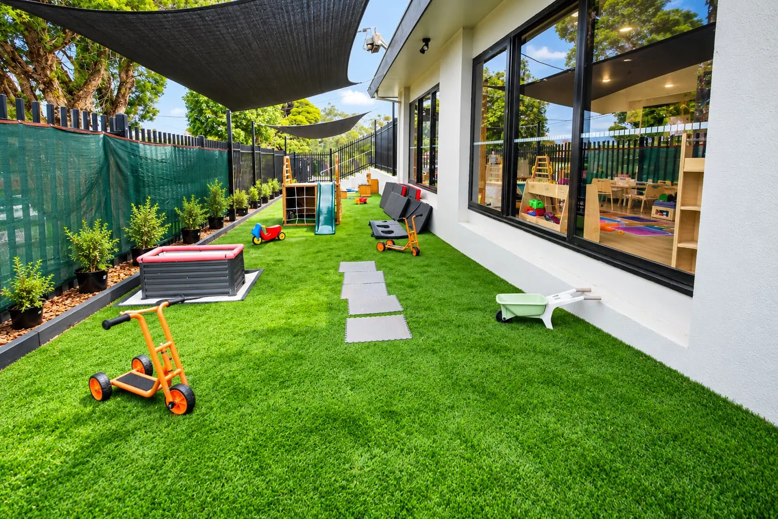 Outdoor play space beside YoMySunshine, with ride-on toys on artificial grass under shade sails