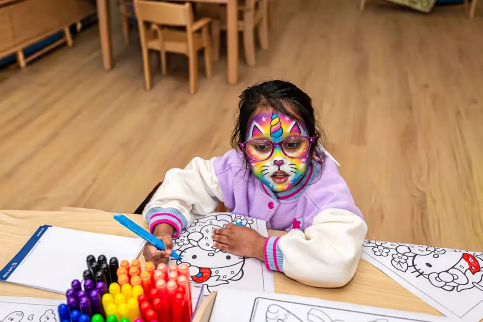 A child with rainbow unicorn face paint colouring in at a table, with a tray of bright markers in front